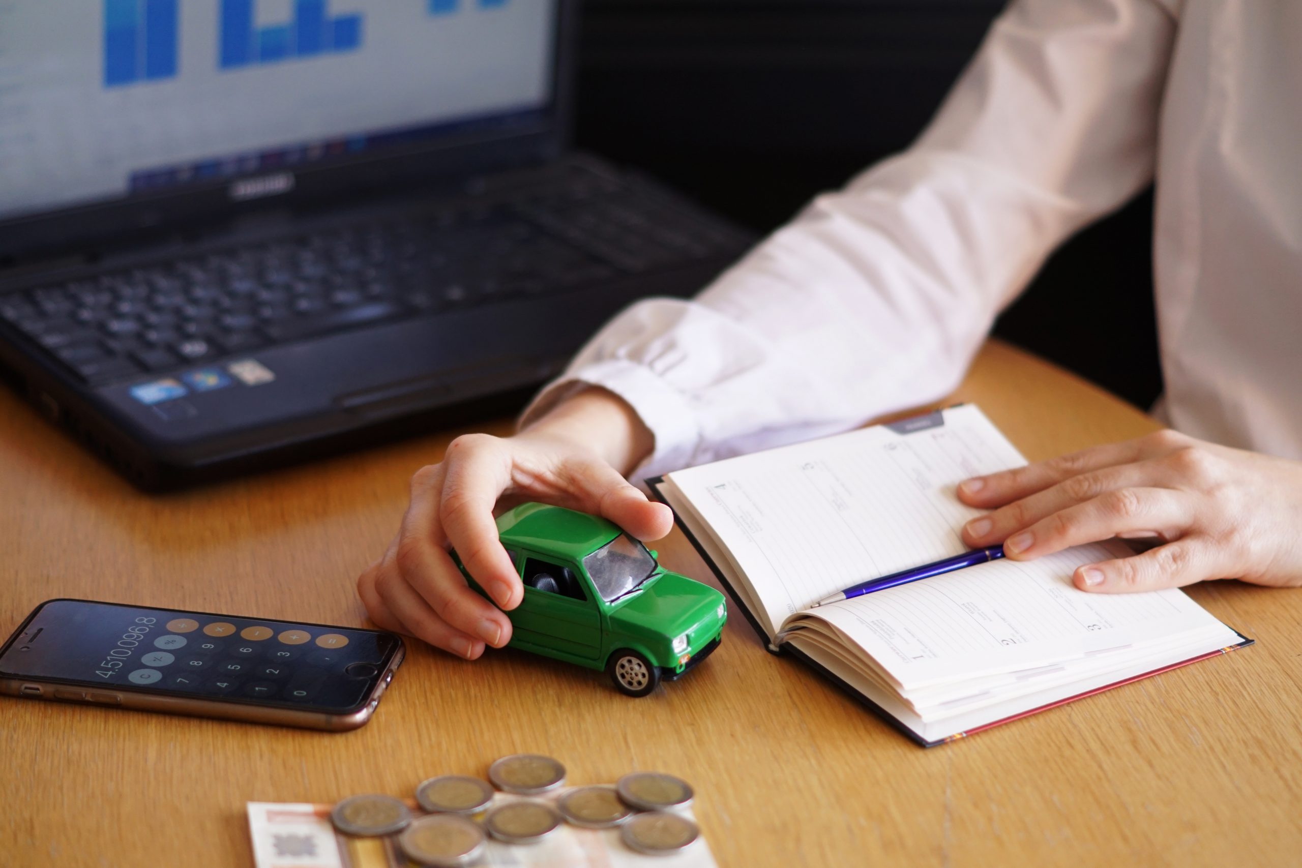 insurance agent holding a green toy car, with an open notebook, a calculator, bills, coins, and a laptop on the desk