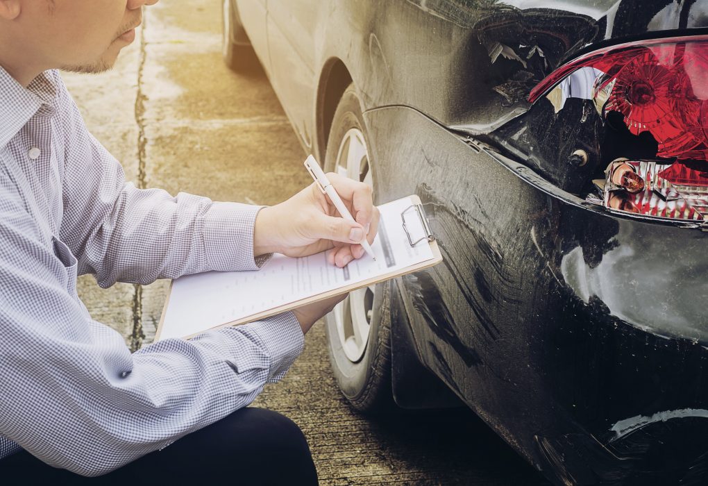 insurance agent writing on a clipboard while inspecting a car's broken tail light