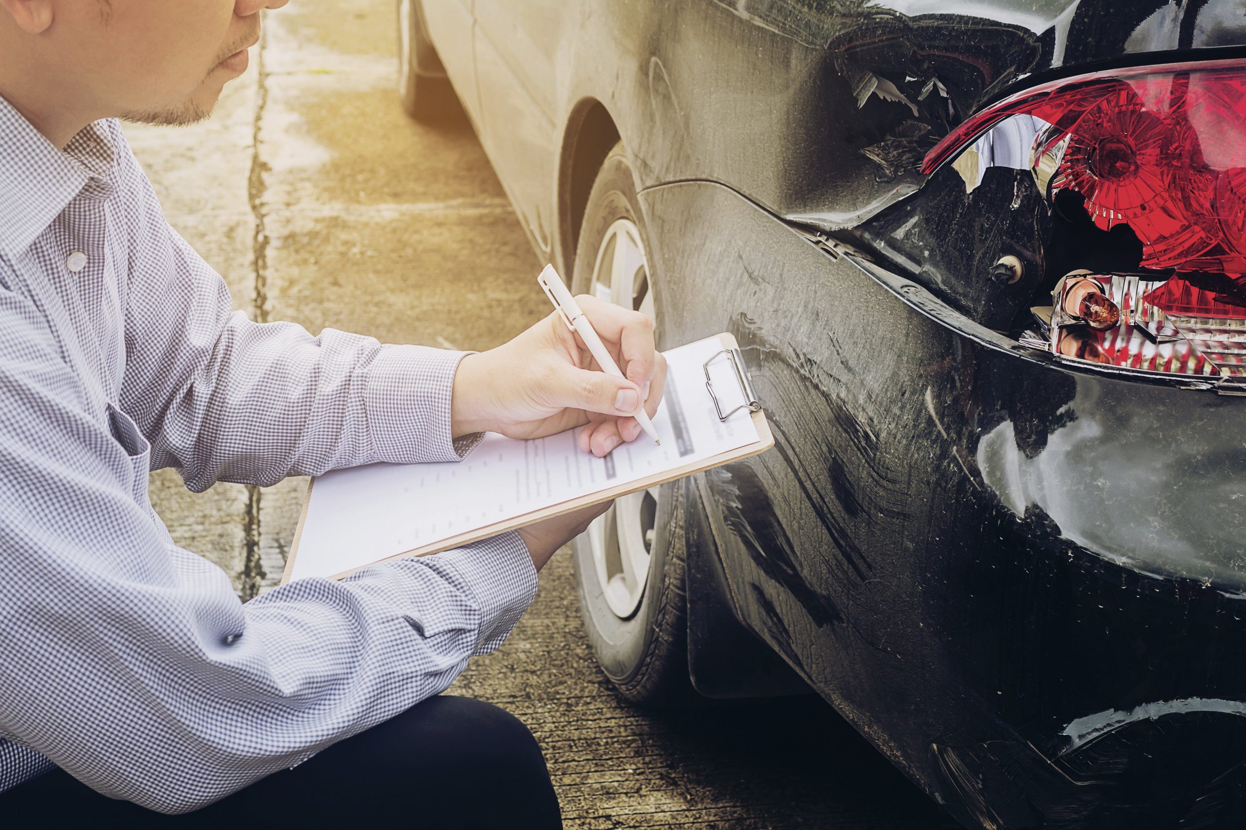 insurance agent writing on a clipboard while inspecting a car's broken tail light